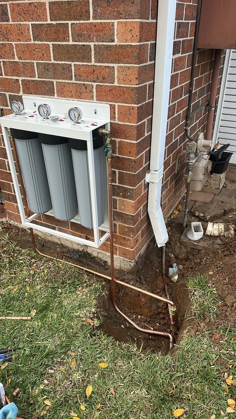 Outdoor water filtration system mounted on brick wall with copper pipes and filter cartridges visible