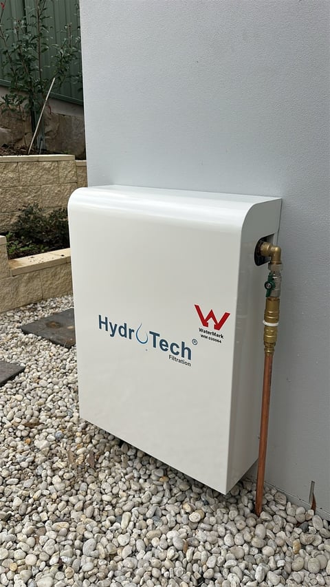 White HydroTech water filter unit mounted on outdoor wall with copper pipe connection and gravel ground