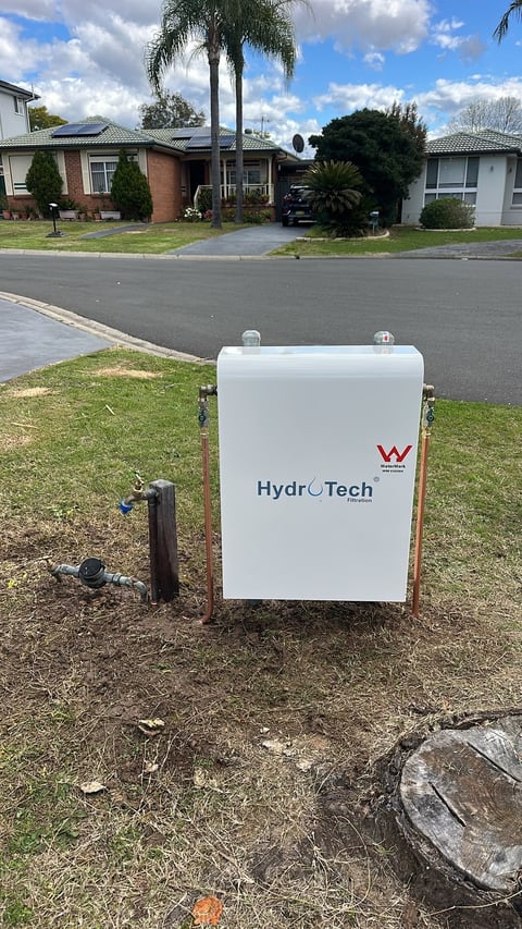 White HydroTech water tank installed on residential street with brick houses and palm tree in background