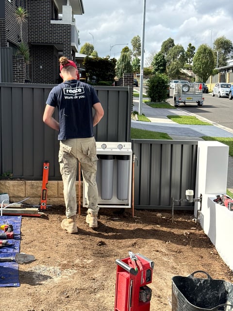 Worker in navy shirt standing on construction site near metal fence and HVAC unit, viewed from behind