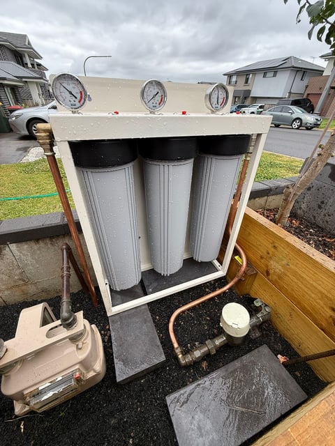 Water filtration system with three cylindrical filters mounted in a beige housing unit, featuring pressure gauges and copper piping on a residential driveway