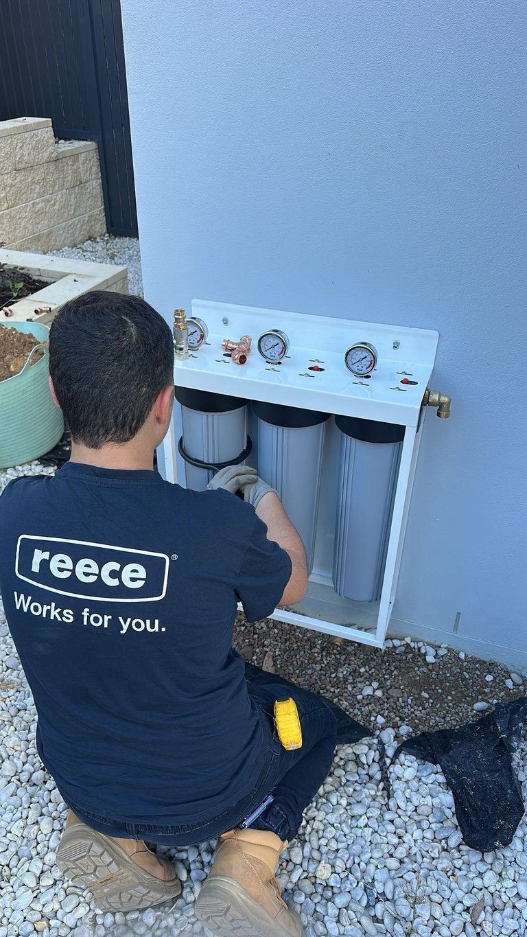 Technician in Reece work shirt installing a white water filtration system with three cylindrical filters against a blue wall