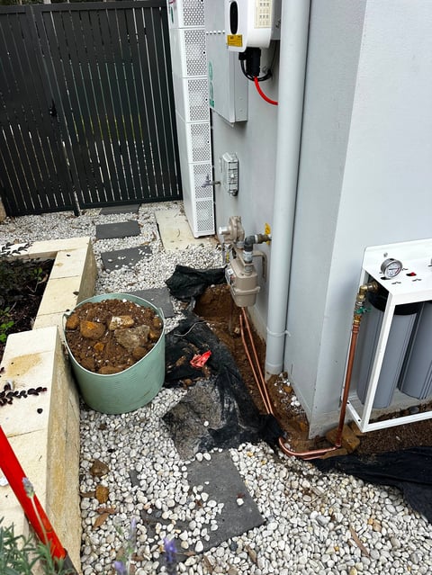 Outdoor utility area with green bucket, black piping, white gravel ground, gray fence, and HVAC equipment against white wall