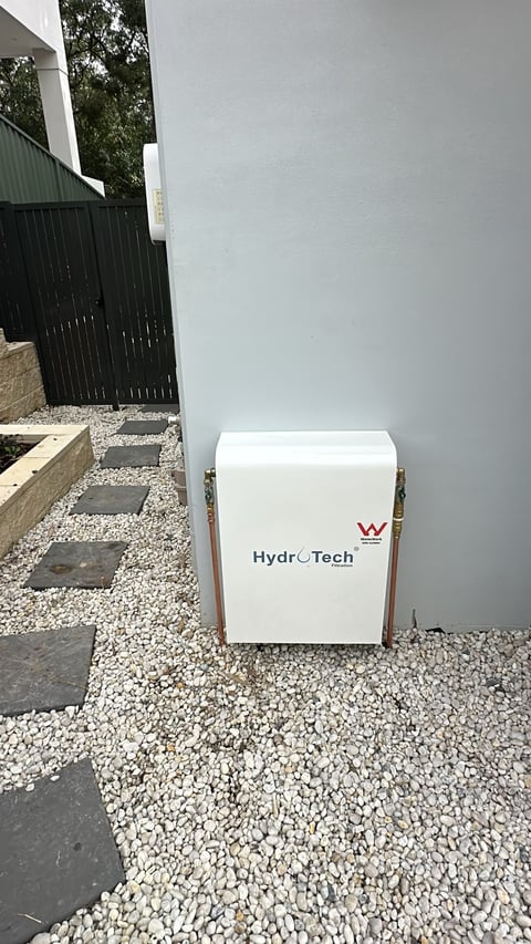 White HydroTech unit installed outdoors against gray wall on white gravel with black gate visible in background
